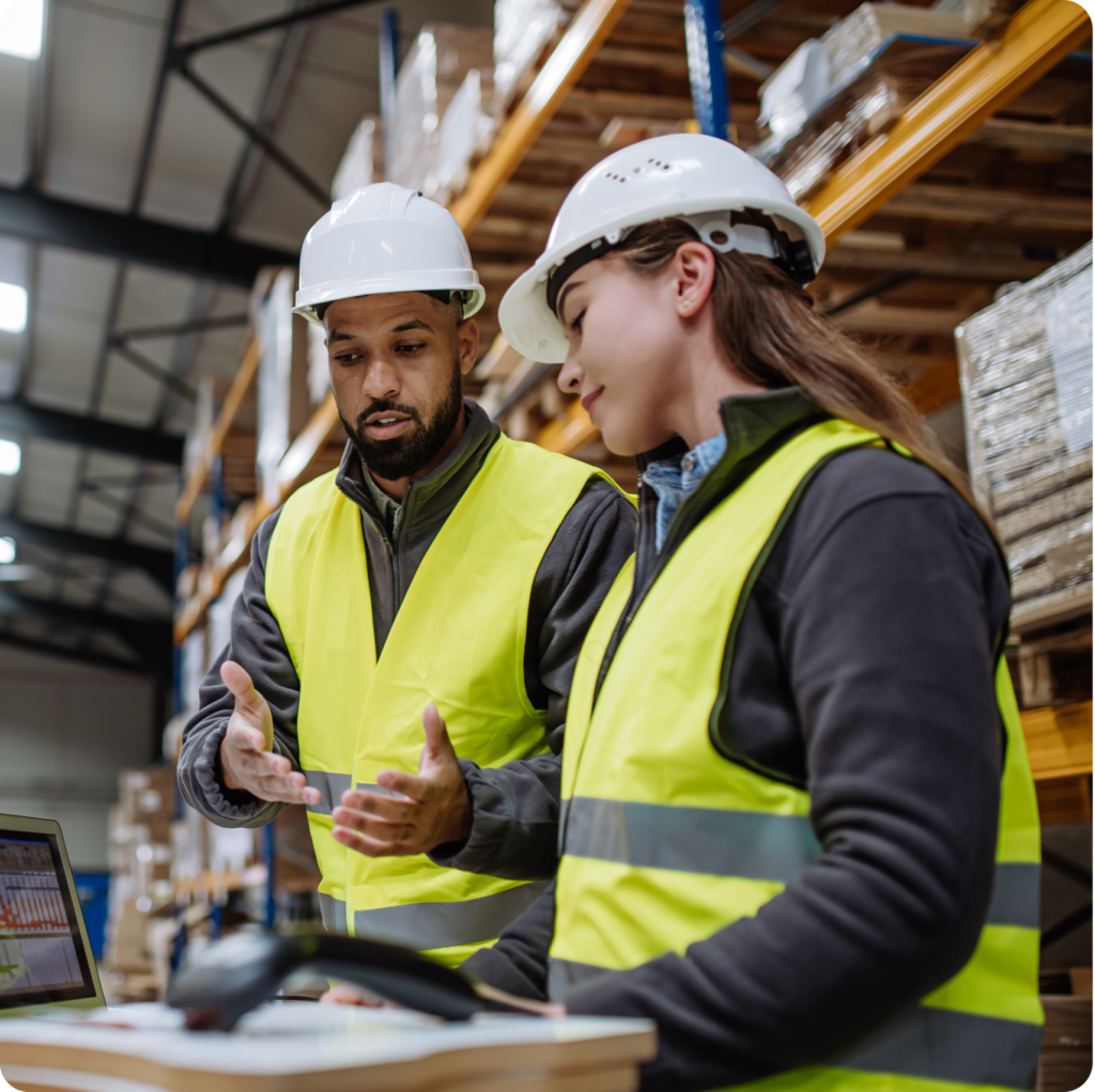 Warehouse team in hard hats and safety vests checking inventory on a laptop in a stocked distribution warehouse